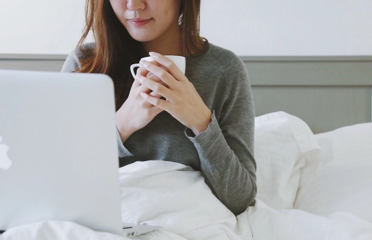 Woman sitting in bed looking at her laptop and cradling a mug