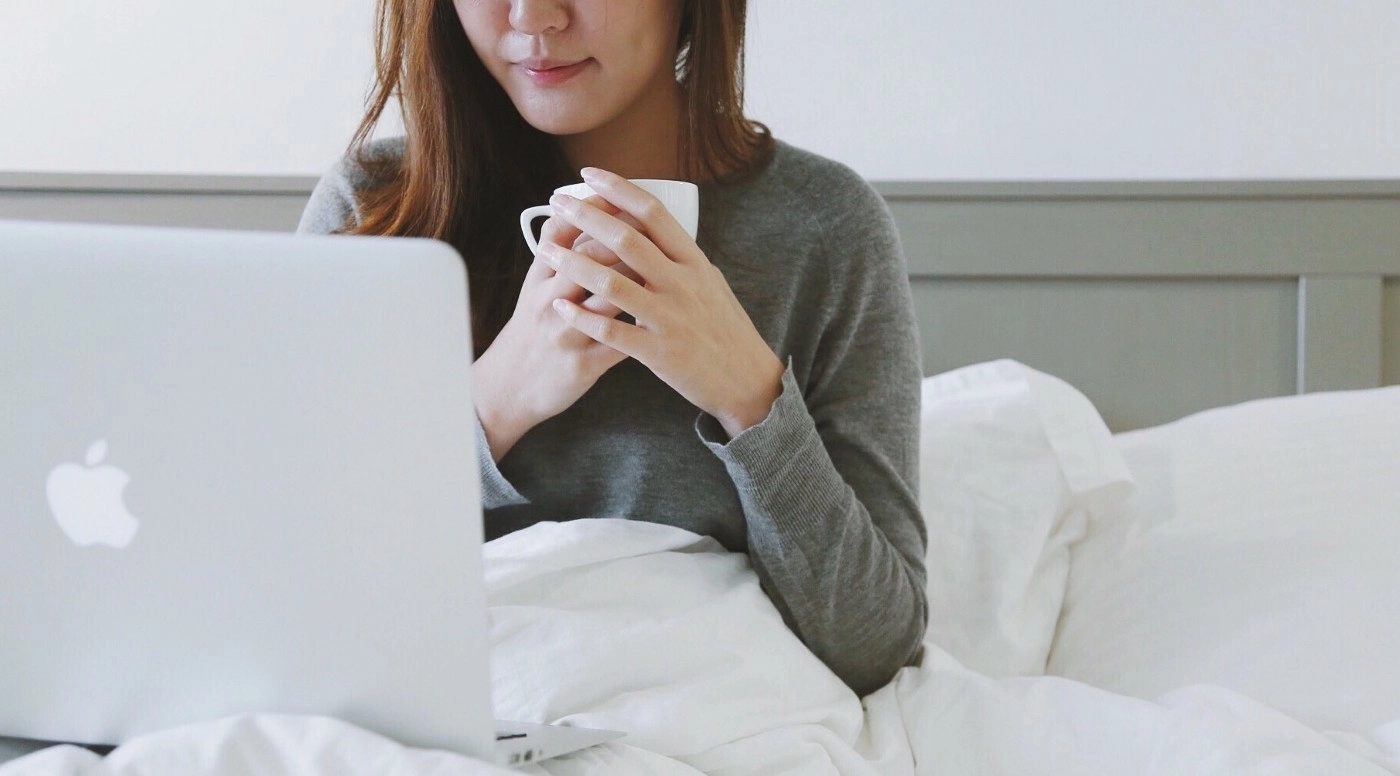 Woman sitting in bed looking at her laptop and cradling a mug