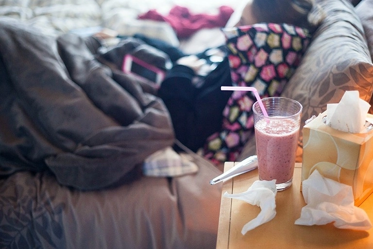 Tissues, a thermometer and a smoothie sitting on a nightstand next to a bed where someone is resting