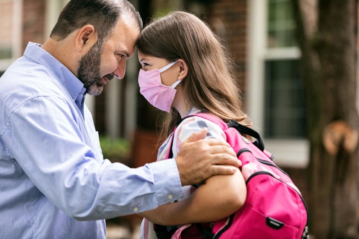 Father and daughter in a warm embrace, as the daughter wears a backpack and a fabric mask