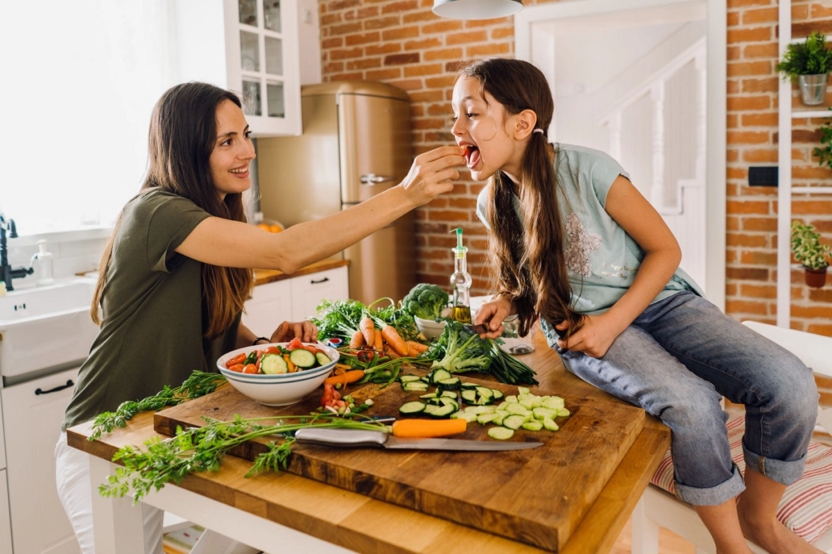 Woman feeding a vegetable to a girl, who is sitting on the kitchen counter next to lots of vegetables