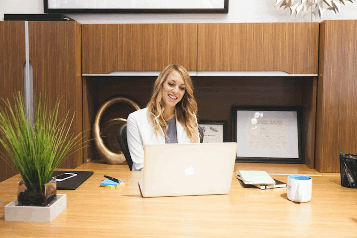 A woman in a white coat sitting at a desk, looking at her laptop and smiling