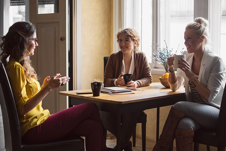 Three women sitting around a table talking and drinking from mugs
