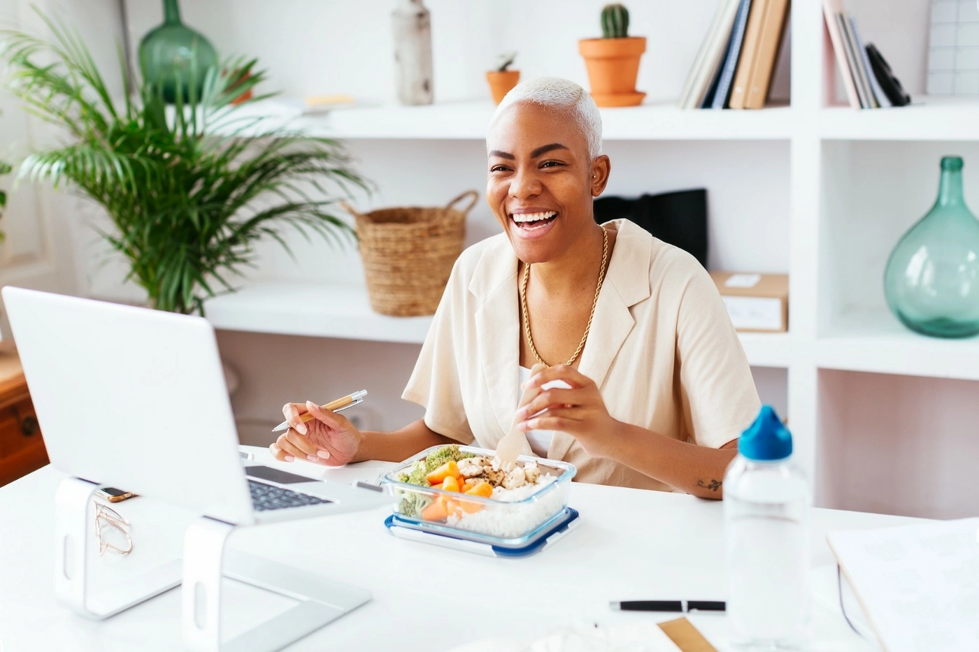 Woman sitting at her desk laughing, eating lunch and looking at her laptop