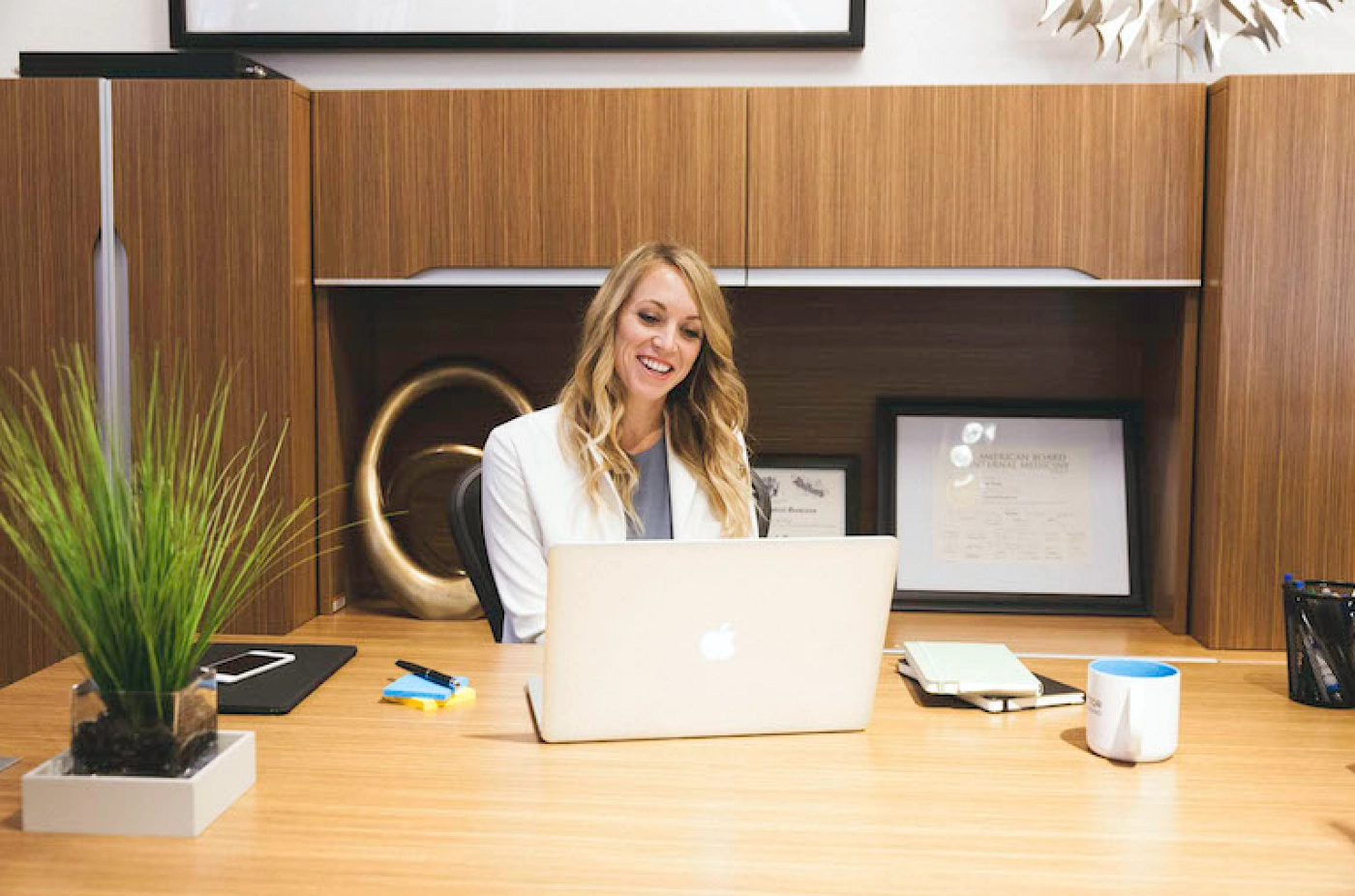 A woman in a white coat sitting at a desk, looking at her laptop and smiling
