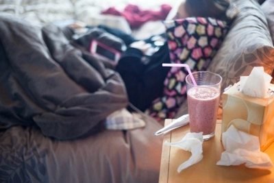 Tissues, a thermometer and a smoothie sitting on a nightstand next to a bed where someone is resting