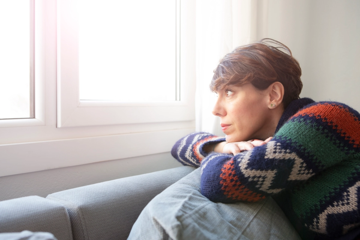 A woman in a sweater sits on a sofa and gazes out the window