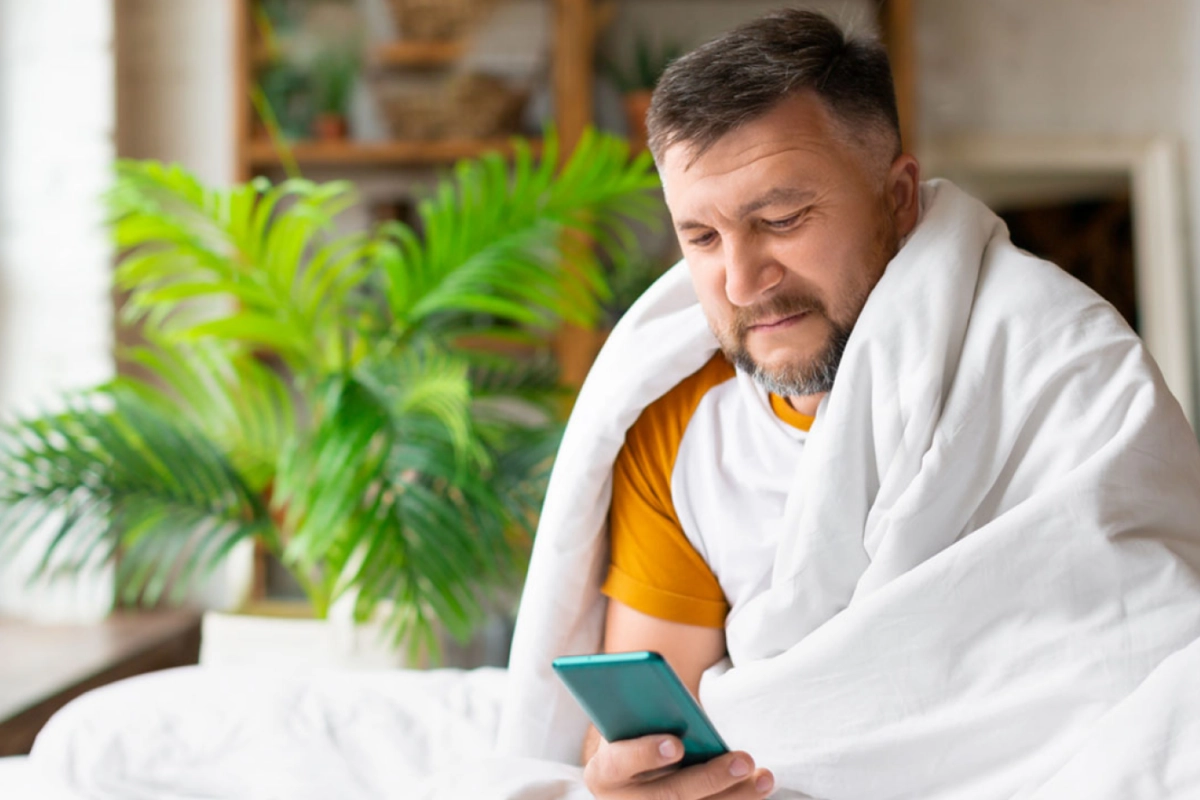 A man, covered in a blanket, looks at his phone while recovering from Covid.