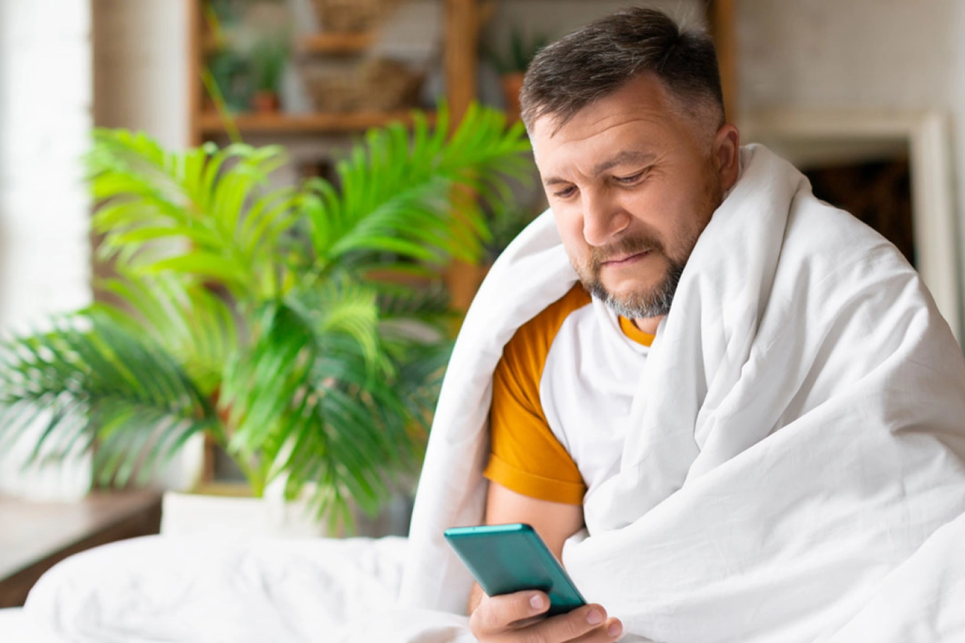 A man, covered in a blanket, looks at his phone while recovering from Covid.