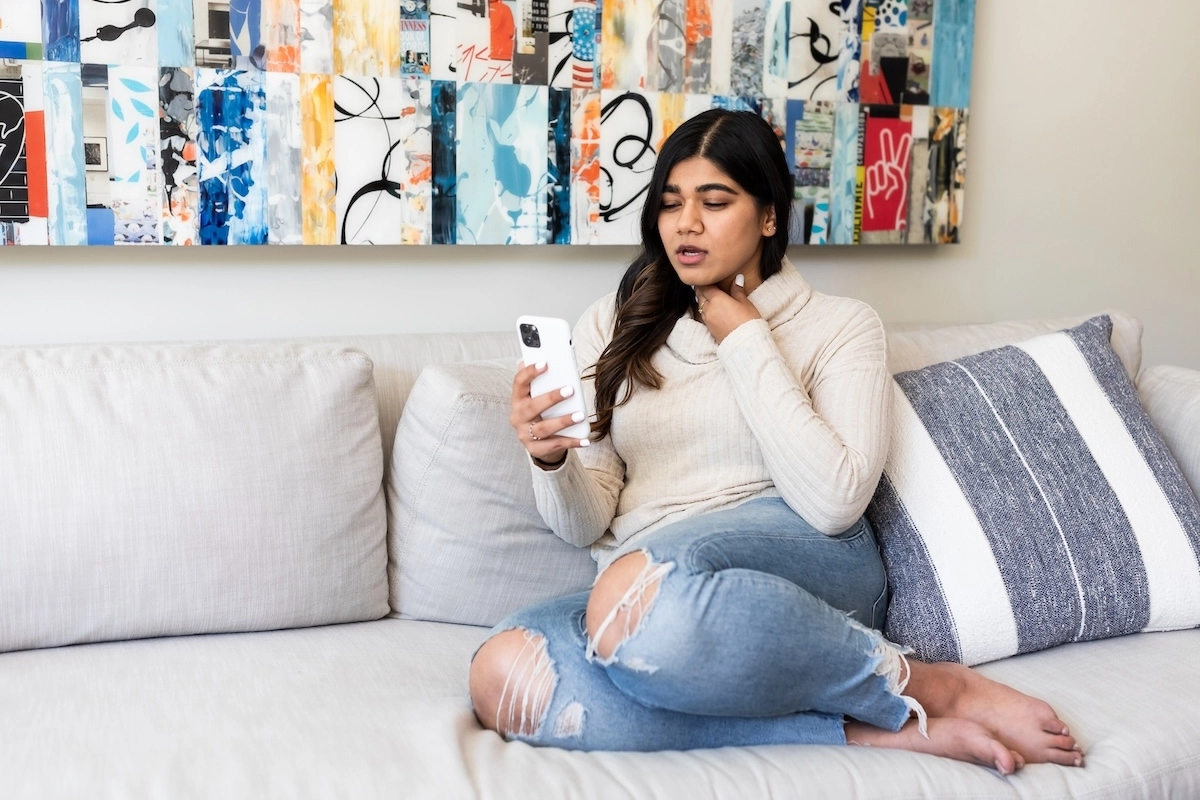 Young woman using Doctor On Demand touching her sore neck sitting on the couch.