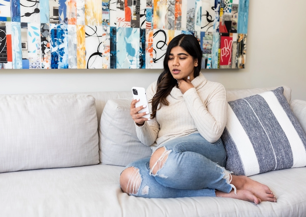 Young woman using Doctor On Demand touching her sore neck sitting on the couch.