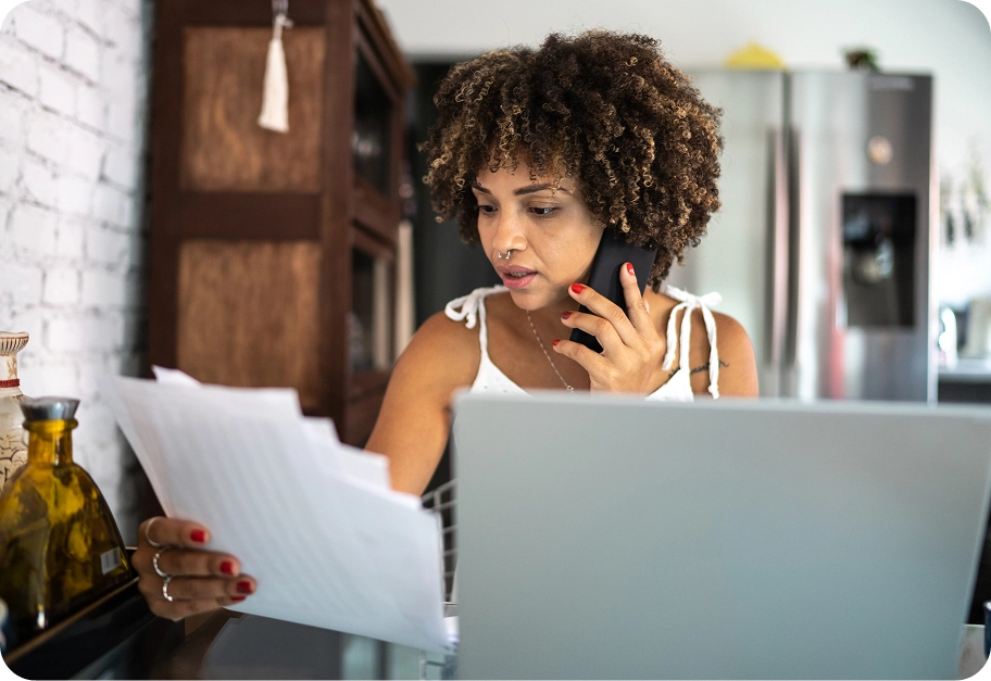 A woman with curly hair sits at a table with a laptop, holding a stack of documents and talking on a mobile phone.