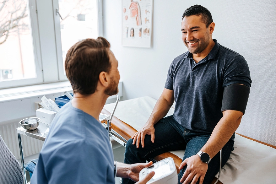 A male doctor in blue scrubs checks the blood pressure of a smiling male patient sitting on an examination table in a clinic room.