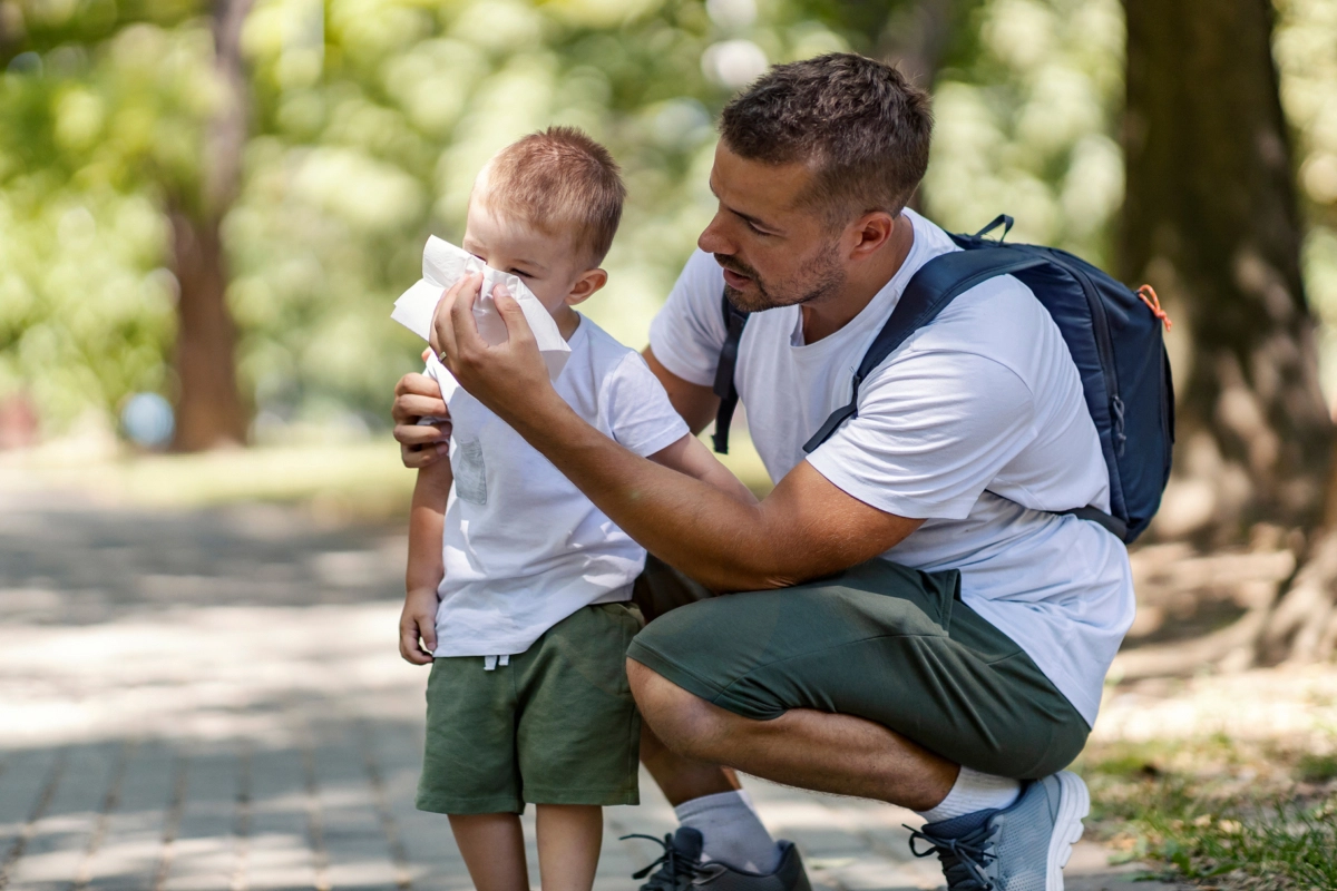A father holding a tissue for his son outside in a park.