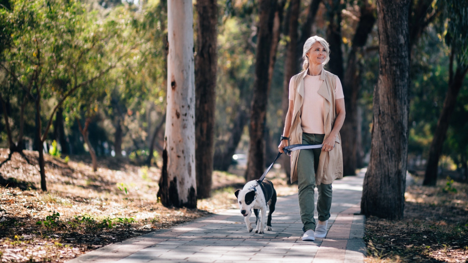 An older woman walking her dog outside in a forest.