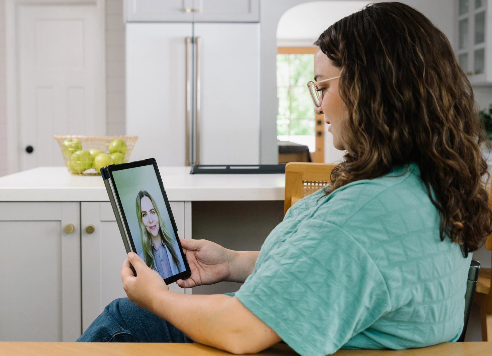 A woman sitting in her kitchen and having a conversation with a medical professional on her iPad.